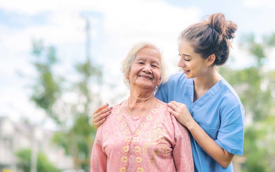 Eine Pflegerin berührt, eine ältere Dame an den Schultern, beide lächeln.